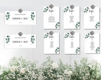 May include: Six white table cards with green eucalyptus leaves and a silver rhinestone embellishment. Each card has a table number and a list of names. The top two cards are larger and say "Please find your seat" and "Kerwin & Alec".