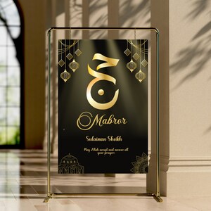 Puede incluir: Un soporte dorado con un letrero negro que dice "Mabror" en caligrafía dorada. El letrero también tiene el nombre "Sulaiman Sheikh" y el texto "Que Allah acepte y responda todas tus oraciones".