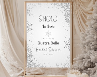 May include: A gold-framed sign with the words "SNOW In Love" and "Welcome to Quatra Belle Bridal Shower 12.20.2025" in elegant script. The sign is decorated with snowflake accents and sits on a white surface.