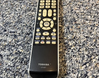 May include: A black and white Toshiba CT-877 remote control with numerous buttons for TV and DVD functions. The remote has a curved bottom and a silver trim. The buttons are beige and black, with numbers, volume, and menu controls.