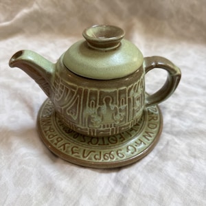 May include: A small ceramic teapot with a matching saucer, featuring a light green and brown glaze. The teapot body and saucer are decorated with embossed geometric patterns and symbols, including text around the saucer rim.