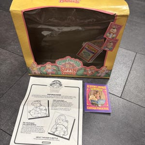 May include: A yellow Cabbage Patch Kids Sippin' Babies toy box with a large rectangular window, accompanied by a white instruction sheet with black illustrations and text, and a small purple booklet titled A Story About My New Baby.