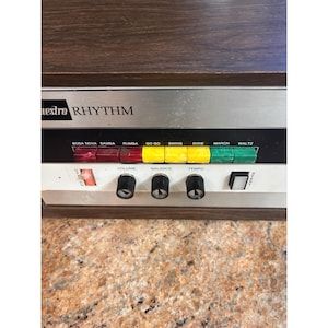 May include: A Maestro Rhythm drum machine with a wood-grain finish, featuring a silver control panel with red, yellow, and green push buttons labeled Bosa Nova, Samba, Rumba, Go Go, Swing, Dixie, March, and Waltz, plus volume, balance, and tempo knobs.