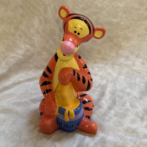 May include: A ceramic figurine of the character Tigger, depicted with orange fur, black stripes, and a pink nose. The character is sitting and holding a blue honey pot with the word HUNNY printed on the front in black, with yellow honey dripping.