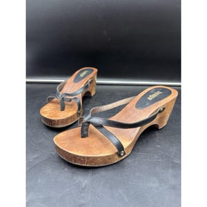 May include: A pair of Bongo thong sandals featuring black leather straps and a medium-toned wooden wedge heel. The footbed is lined with a black oval patch displaying the brand name in gold lettering. The wooden soles show natural grain.