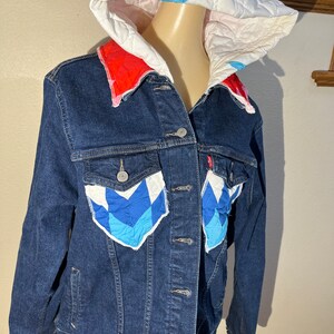 May include: A blue denim jacket with a quilted hood. The hood is lined with white fabric and has a red and white quilted collar. The jacket has two patch pockets on the front, each with a blue and white quilted design.