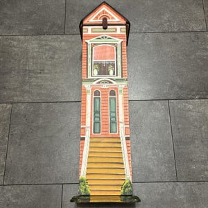 May include: A tall, rectangular wooden wine box featuring a painted facade of a multi-story building with a peaked roof, a central window, two green doors, and a set of stairs leading up to the entrance, finished with a small metal latch.