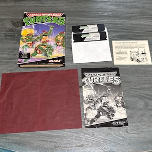 May include: A vintage Teenage Mutant Ninja Turtles video game set. The box features the Turtles with weapons. Includes game cartridges, a manual, and a red sheet. The game is labeled "Ultra".