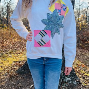 May include: A white sweatshirt with a patchwork design featuring a pink, yellow, and blue star-shaped applique. The sweatshirt has long sleeves and a crew neck.
