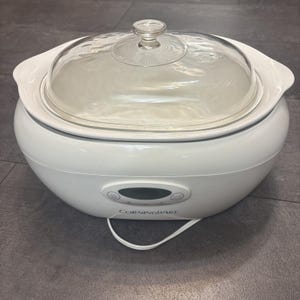 May include: White Corningware slow cooker with a clear glass lid. The slow cooker has a digital display and control buttons. The power cord is white. The slow cooker is designed for cooking meals.
