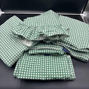 May include: Green and white gingham patterned bedding set. The set includes a fitted sheet, a flat sheet, and pillowcases. One pillowcase has a ruffled edge. The fabric is folded and stacked, showing the pattern clearly.