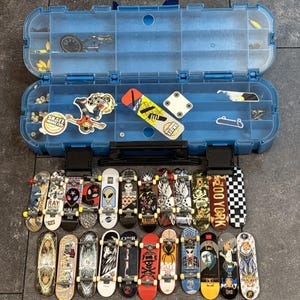 May include: A blue plastic storage case with multiple compartments containing small fingerboard parts, stickers, and a collection of twenty-one miniature toy skateboards with various graphic designs, including skulls, aliens, and logos.