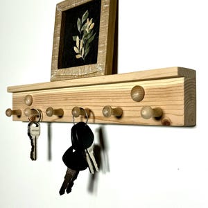 May include: A wooden key rack with six hooks and a small framed picture of a floral design. The key rack is made of light wood and has a natural finish. The hooks are made of wood and have a rounded shape. The picture frame is made of wood and has a distressed finish.
