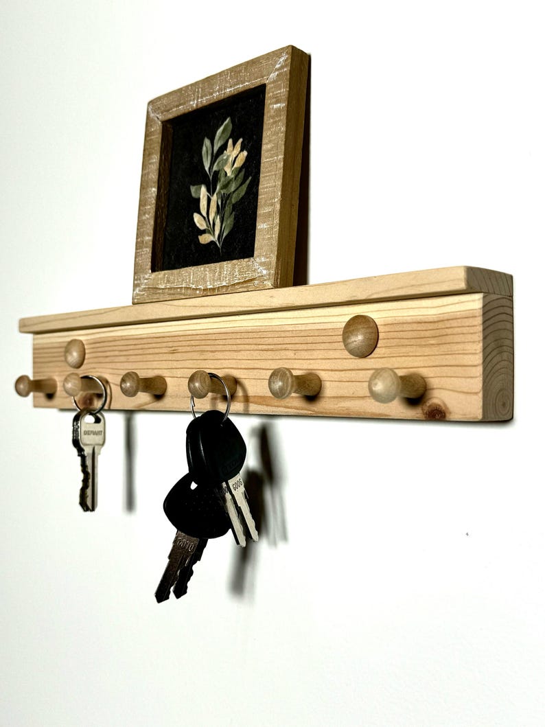 May include: A wooden key rack with six hooks and a small framed picture of a floral design. The key rack is made of light wood and has a natural finish. The hooks are made of wood and have a rounded shape. The picture frame is made of wood and has a distressed finish.