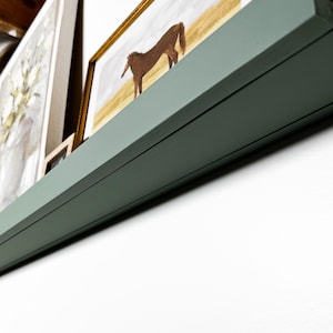 May include: A green wooden shelf with a brown horse painting in a gold frame on top. The shelf is mounted on a white wall.