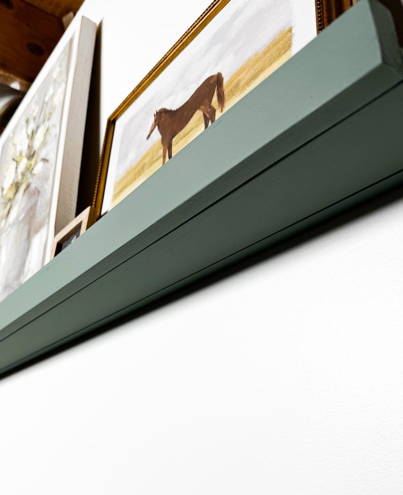 May include: A green wooden shelf with a brown horse painting in a gold frame on top. The shelf is mounted on a white wall.