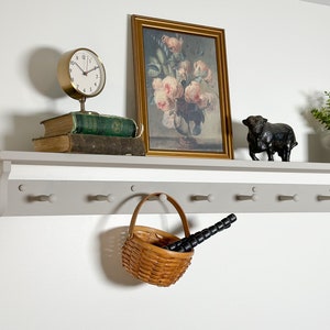 May include: A gray wooden shelf with white knobs and a basket hanging from one of the knobs. The shelf is decorated with a clock, books, a framed floral print, a small black bull figurine, and a potted plant.