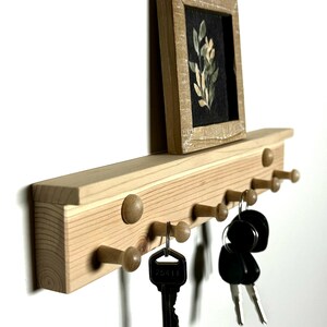 May include: Wooden key rack with a small framed picture of a floral design. The rack has eight hooks and is mounted on a white wall. There are keys hanging on the hooks.