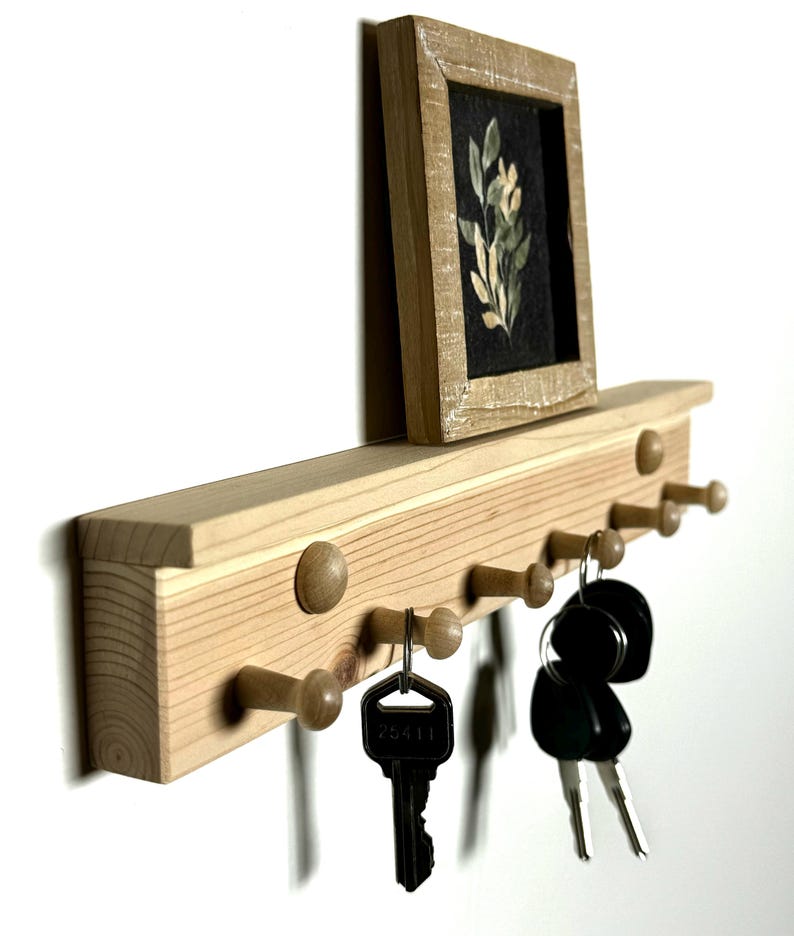 May include: Wooden key rack with a small framed picture of a floral design. The rack has eight hooks and is mounted on a white wall. There are keys hanging on the hooks.