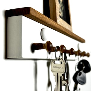 May include: White wooden key rack with a small wooden shelf above it. The shelf has a framed picture on it. The key rack has five hooks with keys hanging on them. The key rack is mounted on a white wall.