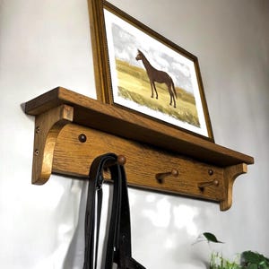 Pode incluir: Uma prateleira de parede de madeira com três ganchos e uma imagem de um cavalo num campo. A prateleira é tingida de uma cor castanho escuro e tem um design simples. Uma correia de couro preto pendura em um dos ganchos.