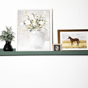 May include: A green shelf with three framed pictures on it. The shelf is mounted on a white wall. The pictures are of a bouquet of flowers, a horse, and a small floral design. A small vase with green leaves and a black base is on the shelf.