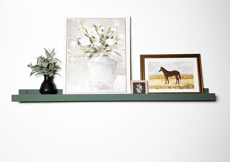 May include: A green shelf with three framed pictures on it. The shelf is mounted on a white wall. The pictures are of a bouquet of flowers, a horse, and a small floral design. A small vase with green leaves and a black base is on the shelf.