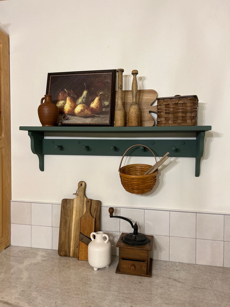 Puede incluir: Una estanter&iacute;a de madera verde con ganchos, una cesta y varios art&iacute;culos de cocina, incluyendo una pintura de peras, un jarr&oacute;n de cer&aacute;mica y un molinillo de caf&eacute; de madera.