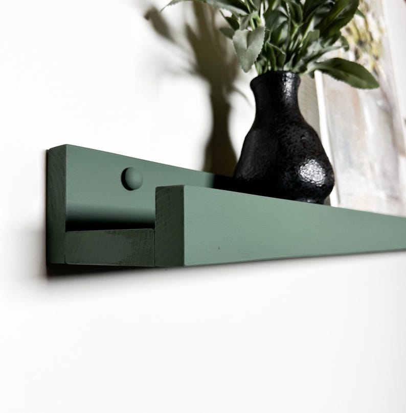 May include: A dark green wooden shelf with a rounded edge and a single screw visible on the front. The shelf is mounted to a white wall.