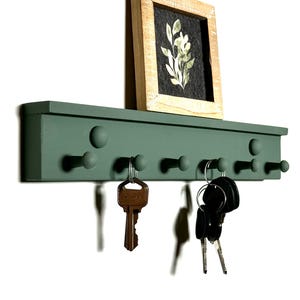 May include: A green wooden key rack with six hooks and a small framed print of a floral design above it. The key rack has a slightly raised edge and rounded knobs on the hooks.