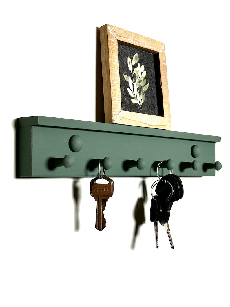 May include: A green wooden key rack with six hooks and a small framed print of a floral design above it. The key rack has a slightly raised edge and rounded knobs on the hooks.