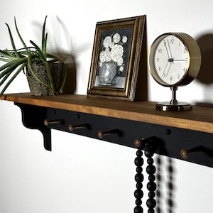 May include: A wooden shelf with a black painted lower section and a natural wood top. It features several bronze-colored hooks and holds a framed picture, a gold-colored clock, and a potted plant. A strand of black beads hangs from a hook.