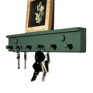 May include: A green wooden key rack with six round knobs for hanging keys. A small framed print with a floral design sits on top of the rack. Several keys are hanging from the knobs.