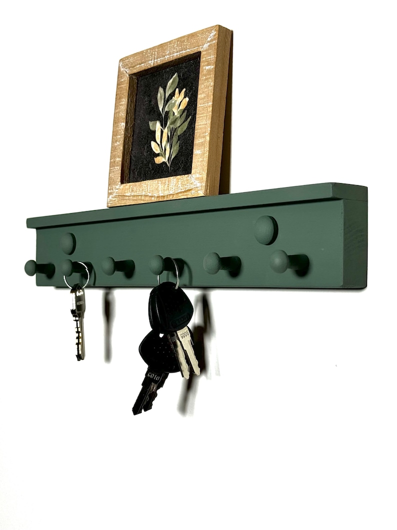 May include: A green wooden key rack with six round knobs for hanging keys. A small framed print with a floral design sits on top of the rack. Several keys are hanging from the knobs.