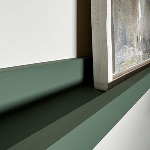 May include: A green painted shelf with a white framed painting on top. The shelf is made of wood and has a simple design.