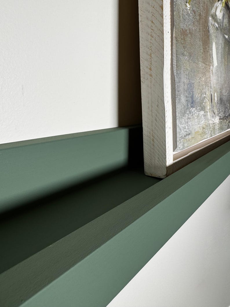 May include: A green painted shelf with a white framed painting on top. The shelf is made of wood and has a simple design.