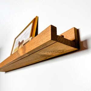 May include: A wooden shelf with a natural finish, mounted on a white wall. The shelf has a unique design with a slight curve and a small lip on the front edge. A framed picture is displayed on the shelf.