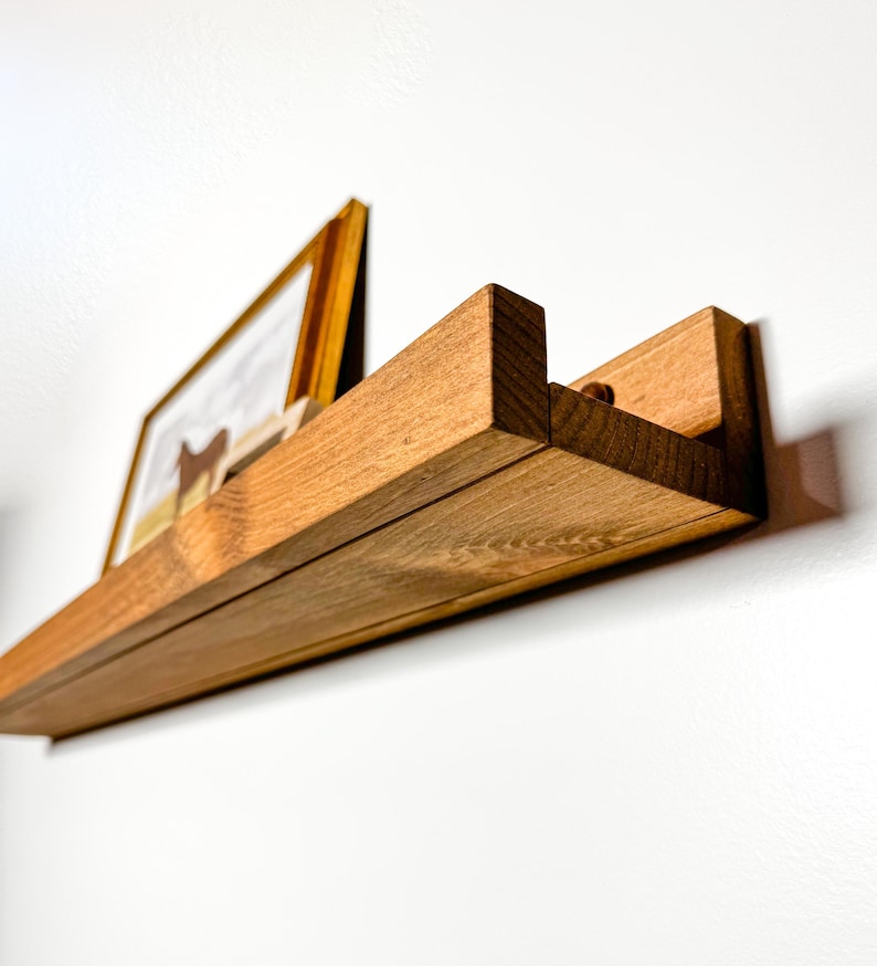 May include: A wooden shelf with a natural finish, mounted on a white wall. The shelf has a unique design with a slight curve and a small lip on the front edge. A framed picture is displayed on the shelf.