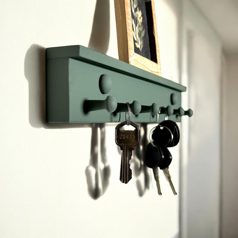 May include: A green wooden key rack with five hooks and round knobs. The rack is mounted on a white wall and has a small framed picture above it. Keys are hanging on the hooks.