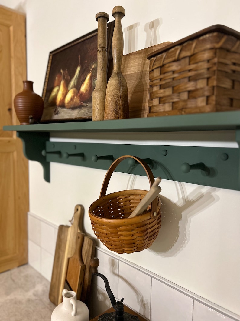 Puede incluir: Una estanter&iacute;a de madera verde con ganchos, una cesta de mimbre marr&oacute;n, un rodillo de madera, un jarr&oacute;n de cer&aacute;mica marr&oacute;n y un cuadro con peras. La estanter&iacute;a est&aacute; montada en una pared de azulejos blancos.