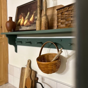 Puede incluir: Una estanter&iacute;a verde con rodillos de amasar de madera, una cesta tejida, una tabla de cortar de madera, una cesta tejida y una pintura de peras. La estanter&iacute;a est&aacute; montada en una pared de azulejos blancos.