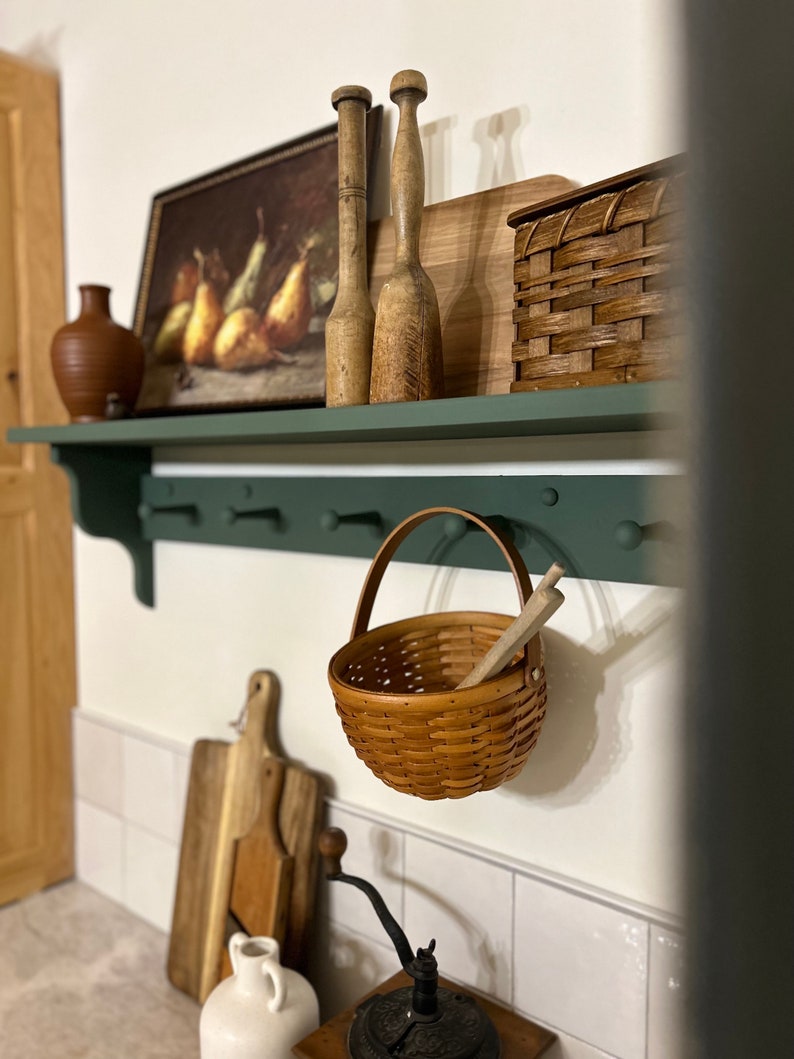 Puede incluir: Una estanter&iacute;a verde con rodillos de amasar de madera, una cesta tejida, una tabla de cortar de madera, una cesta tejida y una pintura de peras. La estanter&iacute;a est&aacute; montada en una pared de azulejos blancos.