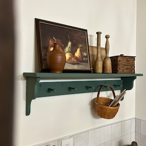 Puede incluir: Un estante pintado de verde con ganchos, una jarra de cer&aacute;mica marr&oacute;n, una pintura enmarcada de peras, clavijas de madera, una cesta de mimbre con tapa y una cesta de mimbre colgando de los ganchos.