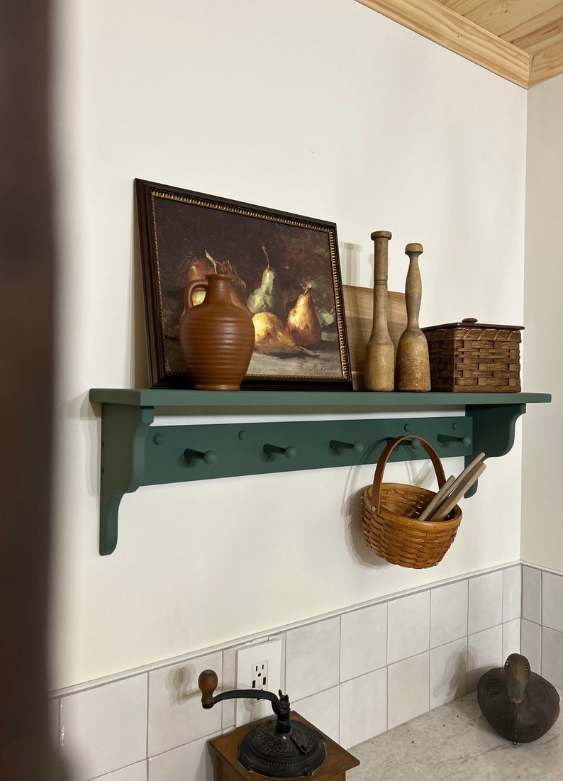 Puede incluir: Un estante pintado de verde con ganchos, una jarra de cer&aacute;mica marr&oacute;n, una pintura enmarcada de peras, clavijas de madera, una cesta de mimbre con tapa y una cesta de mimbre colgando de los ganchos.