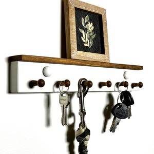 May include: White wooden key rack with a shelf on top. The shelf has a small framed picture of a flower on it. The key rack has six hooks and a metal ring. There are keys and a leash hanging on the hooks.