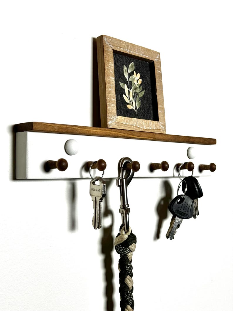 May include: White wooden key rack with a shelf on top. The shelf has a small framed picture of a flower on it. The key rack has six hooks and a metal ring. There are keys and a leash hanging on the hooks.