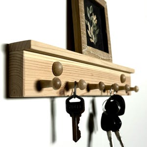 May include: Wooden key rack with a shelf on top. The rack has eight wooden pegs and is painted in a natural wood color. There are keys hanging on the pegs. A small framed picture is on the shelf.