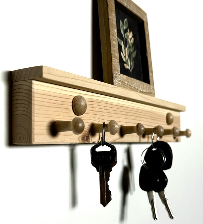 May include: Wooden key rack with a shelf on top. The rack has eight wooden pegs and is painted in a natural wood color. There are keys hanging on the pegs. A small framed picture is on the shelf.