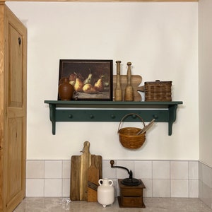 Puede incluir: Un estante verde con una jarra de cer&aacute;mica marr&oacute;n, una pintura enmarcada de peras, clavijas de madera, una cesta de mimbre, una cesta de mimbre marr&oacute;n con asa, una tabla de cortar de madera, una jarra de cer&aacute;mica blanca y un molinillo de caf&eacute; de madera.