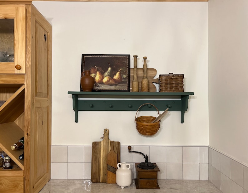 Puede incluir: Un estante verde con una jarra de cer&aacute;mica marr&oacute;n, una pintura enmarcada de peras, clavijas de madera, una cesta de mimbre, una cesta de mimbre marr&oacute;n con asa, una tabla de cortar de madera, una jarra de cer&aacute;mica blanca y un molinillo de caf&eacute; de madera.